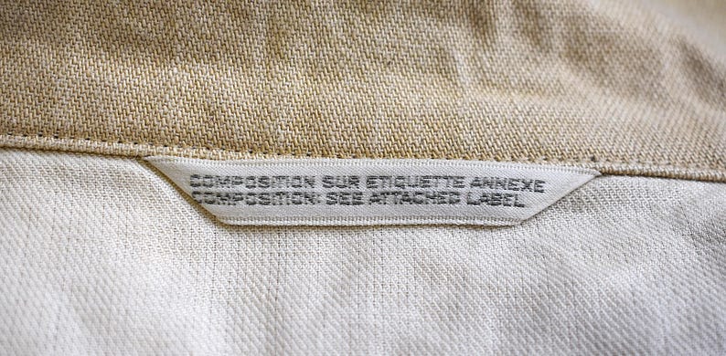 May include: A close-up shot of a garment label, with the text "COMPOSITION SUR ETIQUETTE ANNEXE COMPOSITION: SEE ATTACHED LABEL". The label is white with black text, sewn into the beige and white fabric of the garment.