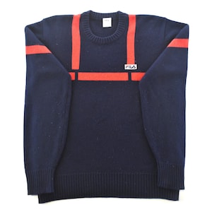 May include: Navy blue sweater with red stripes across the chest and shoulders. The sweater has a crew neck and ribbed cuffs and hem. The Fila logo is on the chest. A classic, casual style.