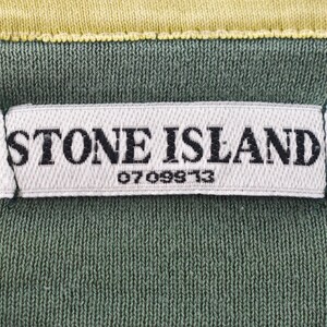 May include: Close-up of a green fabric with a white label that reads "STONE ISLAND" in black capital letters. Below the brand name is the number "0709913". The top edge is yellow.