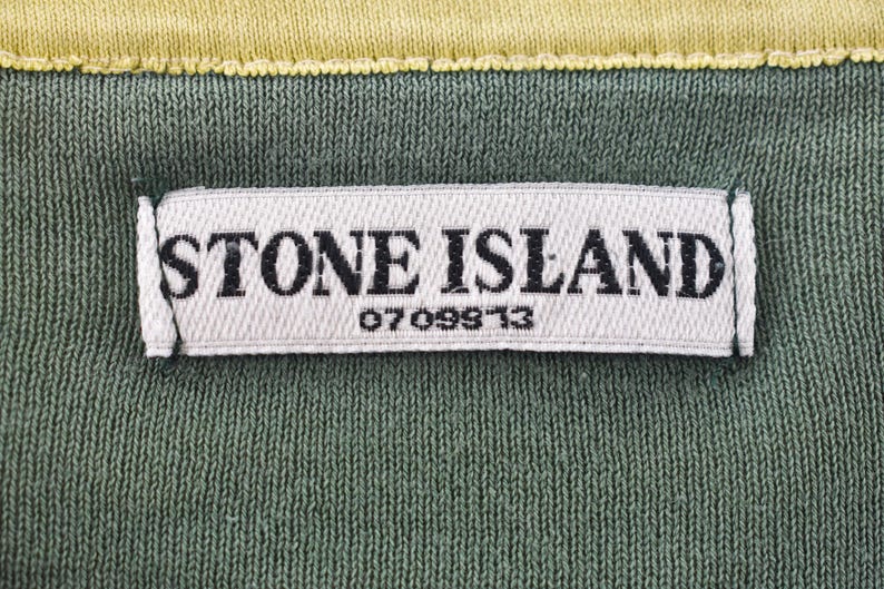 May include: Close-up of a green fabric with a white label that reads "STONE ISLAND" in black capital letters. Below the brand name is the number "0709913". The top edge is yellow.