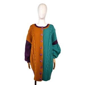 Può includere: Un cardigan oversize a blocchi di colore nei toni dell'arancione, del verde acqua e del viola. Il maglione presenta un motivo a trecce sul lato verde acqua e un'abbottonatura frontale con bottoni viola. Le maniche sono un mix di colori e trame.