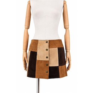 Può includere: Una minigonna in camoscio patchwork caratterizzata da un design a blocchi di colore nelle tonalità marrone scuro, marrone medio e beige. La gonna presenta un orlo dritto e un'abbottonatura verticale frontale con cinque bottoni metallici.