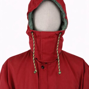 May include: A red hooded jacket with a green-lined hood. The jacket has a high collar, black buttons, and two zippered pockets. The hood has a drawstring with yellow and green accents. The jacket is displayed on a mannequin.