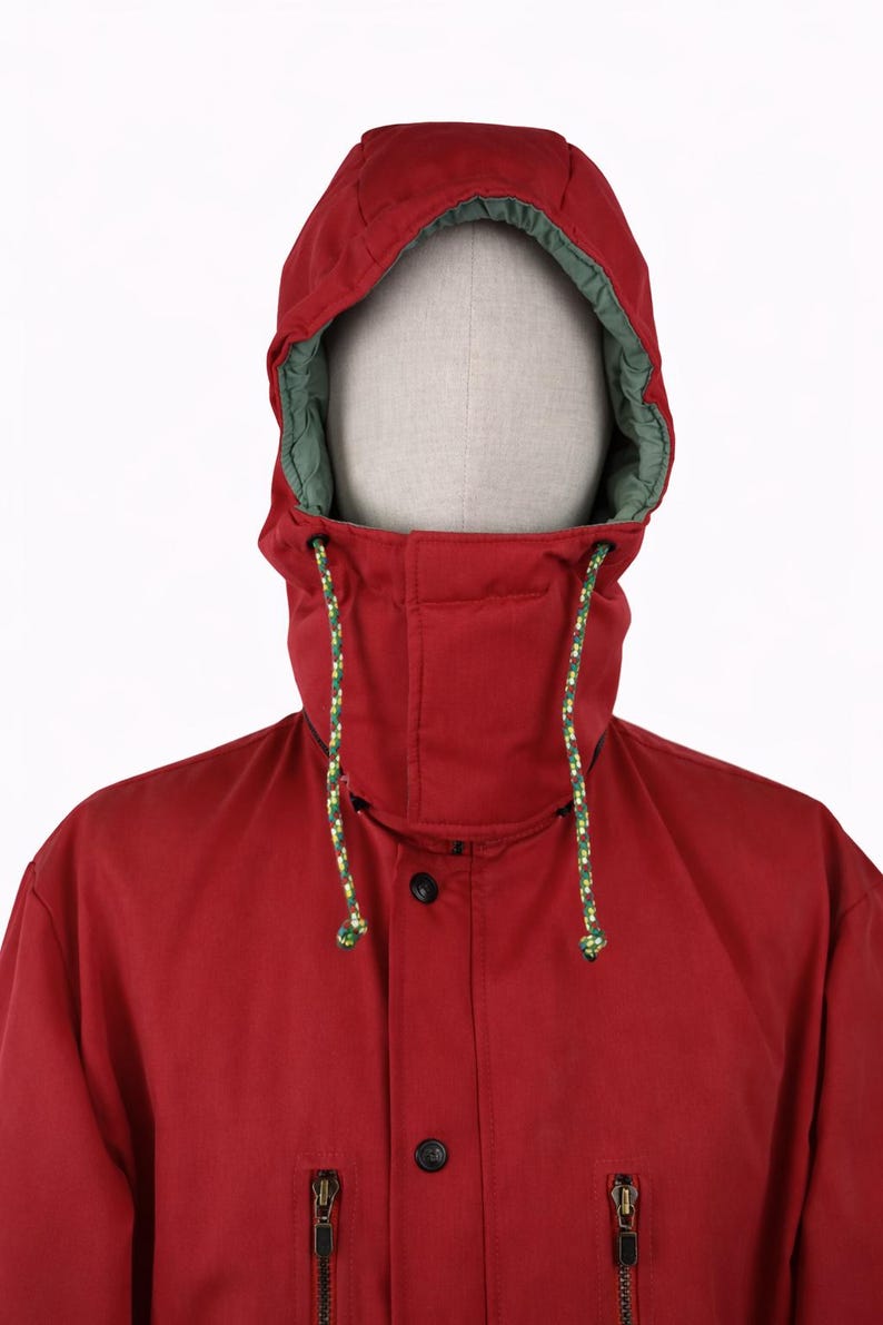 May include: A red hooded jacket with a green-lined hood. The jacket has a high collar, black buttons, and two zippered pockets. The hood has a drawstring with yellow and green accents. The jacket is displayed on a mannequin.