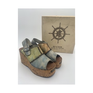 May include: Wedge sandals with a cork sole and a multi-coloured leather upper. The sandals feature a buckle closure and a stitched design. The box behind the sandals has a nautical theme with the words "RIVER LUPO DI MARE".