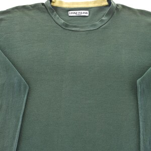 May include: A dark green long-sleeve shirt with a crew neck and a gold-colored neckline. The shirt features a small rectangular label with the text "STONE ISLAND" on the upper chest.