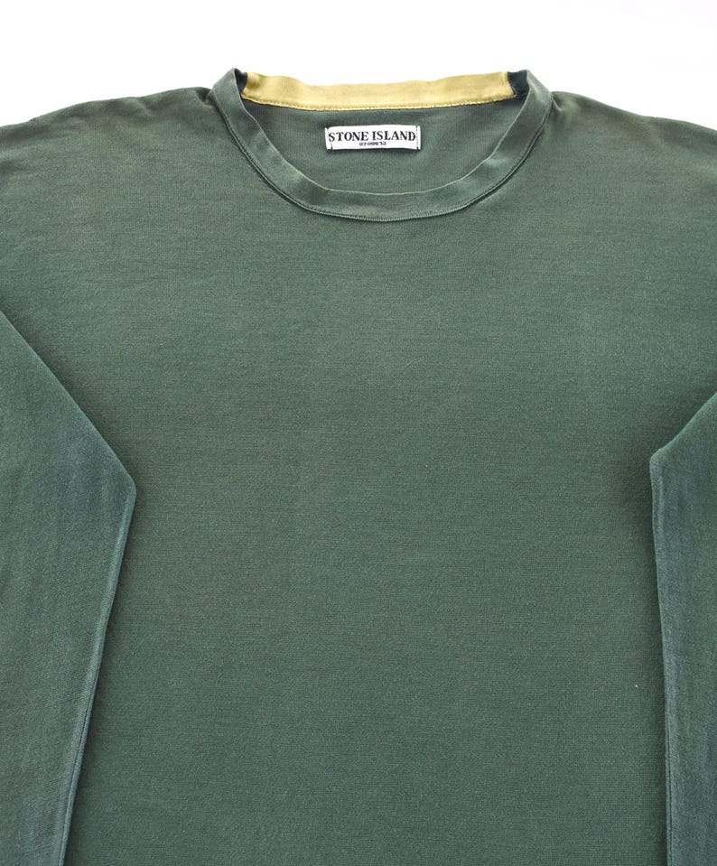 May include: A dark green long-sleeve shirt with a crew neck and a gold-colored neckline. The shirt features a small rectangular label with the text "STONE ISLAND" on the upper chest.
