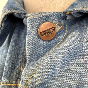 以下が含まれることがあります： 銅色のラングラーボタンが付いたデニムジャケットのクローズアップ。