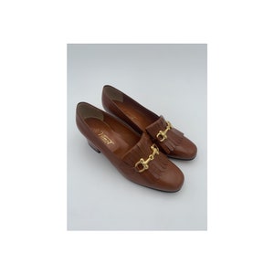 May include: A pair of brown leather heeled loafers with gold metal hardware and fringe detailing. The loafers have a pointed toe and a low heel.
