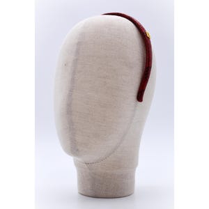 Może przedstawiać: Bordowa opaska do włosów ze wzorem skóry węża i małym, złotym emblematem. Opaska jest prezentowana na głowie manekina na białym tle. To cienka, zakrzywiona opaska.