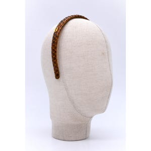 Może przedstawiać: Brązowa opaska do włosów ze skóry węża ze złotą klamrą. Opaska jest prezentowana na głowie manekina. Wzór skóry węża ma odcienie brązu i beżu. Opaska to modny dodatek.