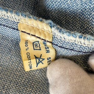 以下が含まれることがあります： 青いデニム生地に縫い付けられた黄色の布製ラベル。ラベルには「Wash Separately」と「100% Cotton」と書かれている。洗濯表示のシンボルが60という数字と共に表示されている。
