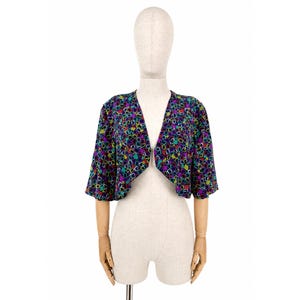 Può includere: Bolero a maniche corte con fantasia floreale colorata. Il capo, presentato su un manichino, presenta uno scollo a V e uno sfondo scuro ornato da forme viola, gialle, verdi e blu.