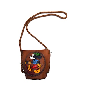 Puede incluir: Un bolso bandolera de cuero marr&oacute;n con un dise&ntilde;o de Mickey Mouse. El bolso tiene una correa trenzada marr&oacute;n y una imagen circular en relieve de Mickey Mouse con su atuendo cl&aacute;sico. El texto "Mickey Mouse" es visible.