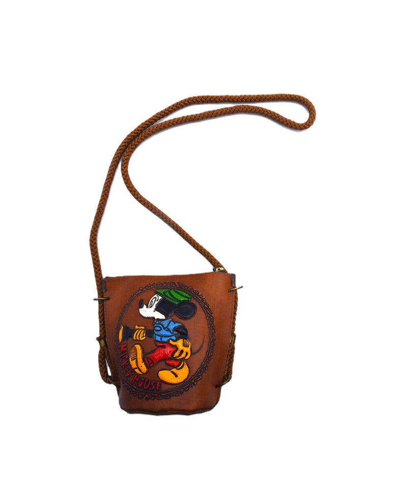 Puede incluir: Un bolso bandolera de cuero marr&oacute;n con un dise&ntilde;o de Mickey Mouse. El bolso tiene una correa trenzada marr&oacute;n y una imagen circular en relieve de Mickey Mouse con su atuendo cl&aacute;sico. El texto "Mickey Mouse" es visible.
