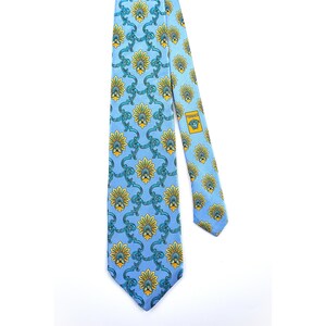 May include: A light blue necktie featuring a repeating pattern of yellow and green floral motifs connected by teal scrollwork. The tie includes a rectangular label on the back with the text Versace and a Medusa head logo in yellow and black.