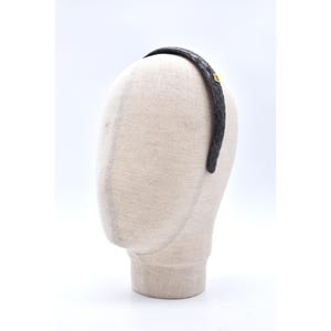 Może przedstawiać: Ciemnoszara opaska do włosów ze wzorem skóry węża jest prezentowana na głowie manekina. Opaska ma cienki, zakrzywiony kształt i mały, złoty emblemat z boku. Tło jest jednolicie białe.