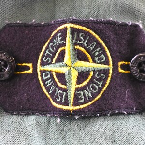 May include: A dark purple patch with the words "STONE ISLAND" encircling a yellow and green compass rose design. Two black buttons are attached to the patch. The patch is on a green fabric.