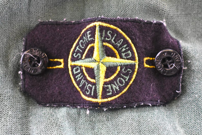May include: A dark purple patch with the words "STONE ISLAND" encircling a yellow and green compass rose design. Two black buttons are attached to the patch. The patch is on a green fabric.