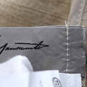May include: Close-up of a grey fabric label with black cursive text. The label is stitched onto a beige fabric. A white fabric is visible in the foreground.
