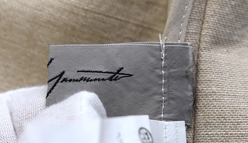 May include: Close-up of a grey fabric label with black cursive text. The label is stitched onto a beige fabric. A white fabric is visible in the foreground.