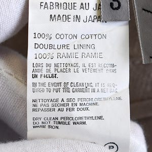 May include: A close-up shot of a garment label with text in French and English. The label states "FABRIQUE AU JA MADE IN JAPA" and provides fabric details and care instructions. A size "3" tag is present.