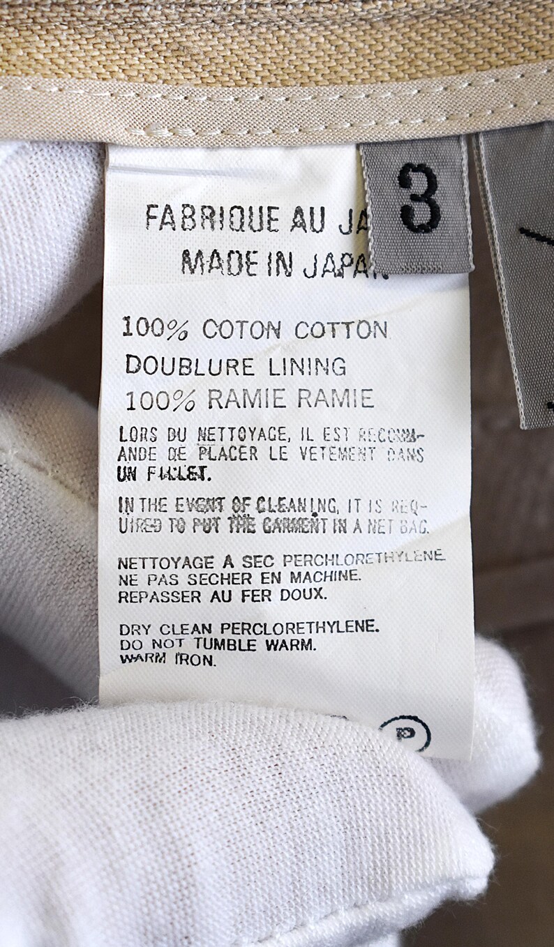 May include: A close-up shot of a garment label with text in French and English. The label states "FABRIQUE AU JA MADE IN JAPA" and provides fabric details and care instructions. A size "3" tag is present.