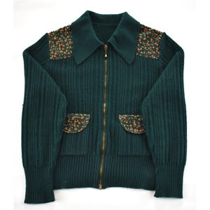 May include: A dark green zip-up cardigan with a ribbed knit texture. The collar and shoulder patches feature a floral pattern with orange and brown accents. The cardigan has two front pockets and a gold-colored zipper.
