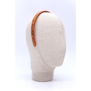 Może przedstawiać: Opaska do włosów ze wzorem skóry węża w odcieniach pomarańczu i brązu. Opaska jest prezentowana na głowie manekina, ukazując jej zakrzywiony kształt i fakturę. Mały, złoty akcent jest widoczny z boku.