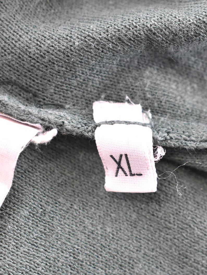 May include: Close-up of a dark green fabric with a pink tag. The tag is white with the letters "XL" printed in black. The fabric appears to be a knit or woven material.