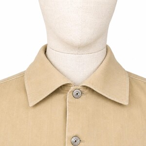 May include: A close-up shot of a tan button-down shirt. The shirt has a classic collar and two silver buttons. The fabric looks like a sturdy cotton or similar material. The shirt is displayed on a mannequin.