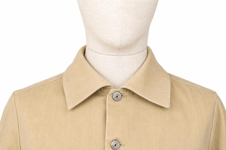 May include: A close-up shot of a tan button-down shirt. The shirt has a classic collar and two silver buttons. The fabric looks like a sturdy cotton or similar material. The shirt is displayed on a mannequin.