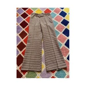 Può includere: Pantaloni a zampa marroni e bianchi con motivo, con chiusura a bottone e passanti per cintura. I pantaloni hanno una vita alta e una gamba svasata. Il tessuto presenta un piccolo disegno geometrico. I pantaloni sono esposti su una coperta colorata a fantasia.