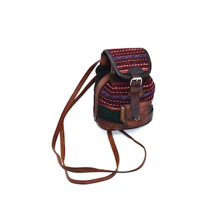 May include: A small backpack with a flap closure and adjustable leather straps. The top flap has a woven design with red, blue, and purple stripes. The body of the bag is a combination of brown leather and woven fabric, with a small front pocket.