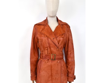 May include: A vintage, rust-coloured leather trench coat on a mannequin. The coat has a double-breasted design, a belted waist with a gold-tone buckle, and buttoned cuffs. The coat's length extends to the mid-thigh.