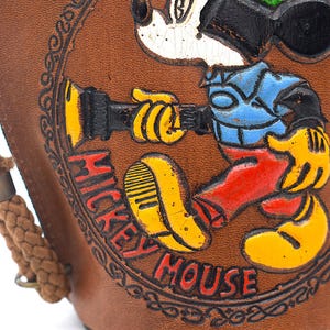 Puede incluir: Art&iacute;culo de cuero marr&oacute;n con una imagen en relieve y colorida de Mickey Mouse. Mickey lleva un sombrero verde, una camisa azul, pantalones rojos y zapatos amarillos. Las palabras "Mickey Mouse" est&aacute;n en rojo.