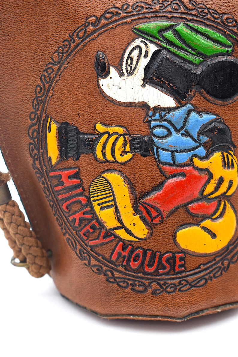 Puede incluir: Art&iacute;culo de cuero marr&oacute;n con una imagen en relieve y colorida de Mickey Mouse. Mickey lleva un sombrero verde, una camisa azul, pantalones rojos y zapatos amarillos. Las palabras "Mickey Mouse" est&aacute;n en rojo.