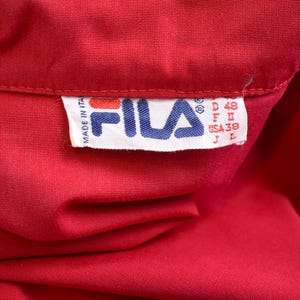 May include: Close-up of a vibrant red garment, possibly a shirt or jacket, with a tag featuring the FILA logo in blue and white. The tag includes size information: D 48, F II, USA 38, and J L. The tag also indicates the item was made in Italy.