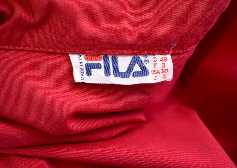May include: Close-up of a vibrant red garment, possibly a shirt or jacket, with a tag featuring the FILA logo in blue and white. The tag includes size information: D 48, F II, USA 38, and J L. The tag also indicates the item was made in Italy.