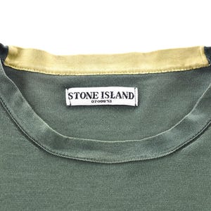 May include: Close-up of a green t-shirt with a contrasting tan collar. The shirt features a tag with the text "STONE ISLAND" and the number "0709913". The fabric appears to be a soft, knit material.