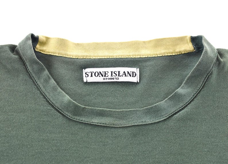 May include: Close-up of a green t-shirt with a contrasting tan collar. The shirt features a tag with the text "STONE ISLAND" and the number "0709913". The fabric appears to be a soft, knit material.