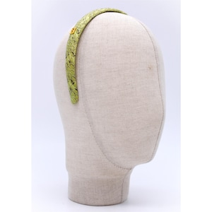 Może przedstawiać: Zielona opaska do włosów ze wzorem łusek i małym złotym akcentem. Opaska jest prezentowana na głowie manekina. Wzór łusek to mieszanka jasnej i ciemnej zieleni. Złoty akcent znajduje się z boku opaski.