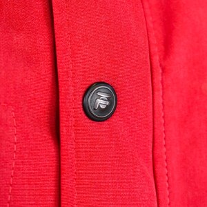 May include: Close-up of a vibrant red fabric with a black button. The button has a silver emblem in the center. The fabric is stitched along the seams.