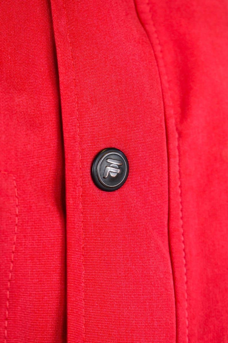 May include: Close-up of a vibrant red fabric with a black button. The button has a silver emblem in the center. The fabric is stitched along the seams.