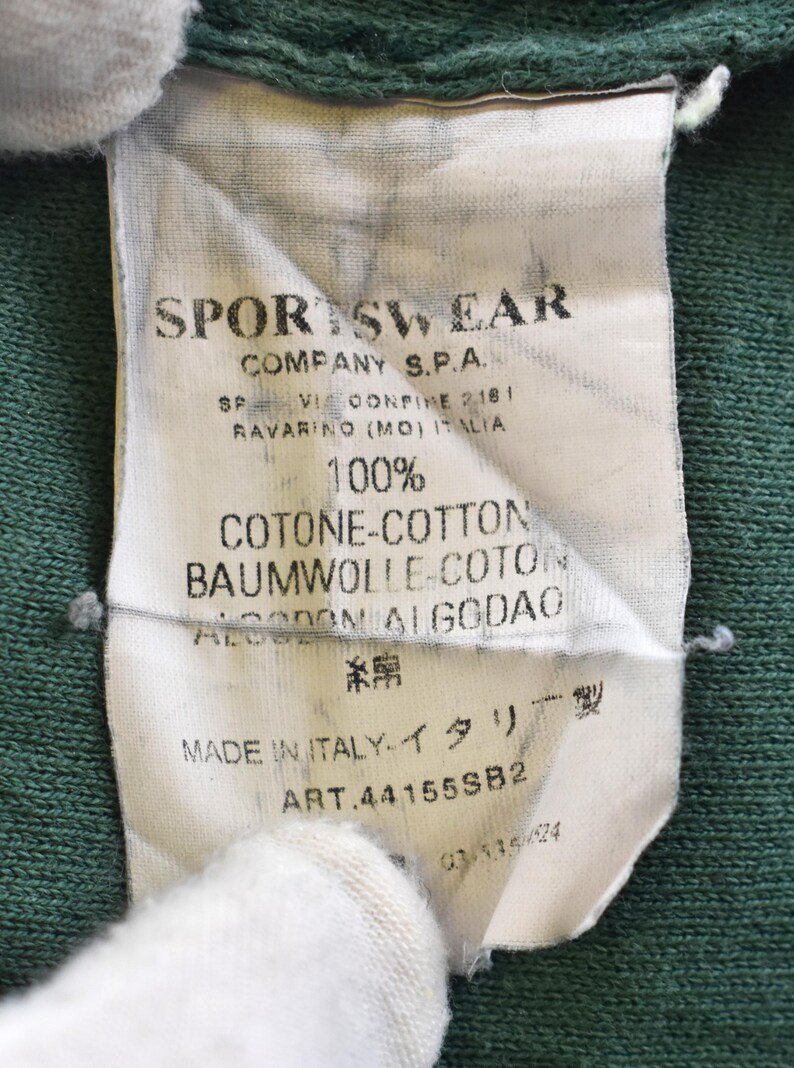 May include: Close-up of a white tag with black text, attached to a green fabric. The tag reads "SPORTSWEAR COMPANY S.P.A." and includes details about the fabric composition, including "100% COTONE-COTTON". The tag also states "MADE IN ITALY."