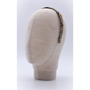 Może przedstawiać: Opaska do włosów z nadrukiem węża jest prezentowana na głowie manekina. Opaska ma wzór czarny, brązowy i beżowy, z małym złotym emblematem. Akcesorium jest przeznaczone do noszenia wokół głowy.