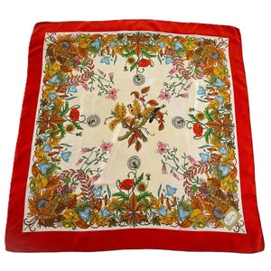 May include: Square silk scarf with a cream-colored center and a vibrant floral and leaf pattern in shades of orange, yellow, blue, and pink. The scarf has a bold red border. A small tag with the brand name is visible.