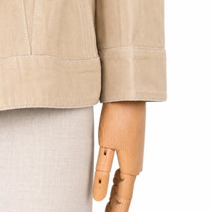 May include: Close-up of a beige jacket with white stitching. The jacket features a structured design and a folded cuff on the sleeve. A wooden hand mannequin is also visible.