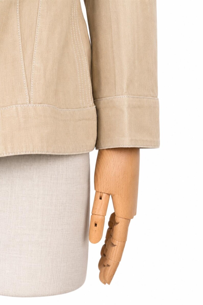 May include: Close-up of a beige jacket with white stitching. The jacket features a structured design and a folded cuff on the sleeve. A wooden hand mannequin is also visible.