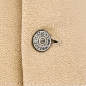May include: Close-up of a beige jacket featuring a silver button. The button is circular and has the text "YAMAMOTO JEANS" around the edge. The jacket's fabric is a plain beige with a visible weave.